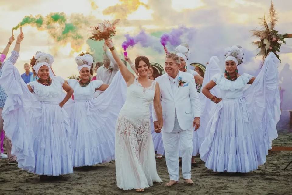 Novios caminando y detrás bailarinas con humo de colores