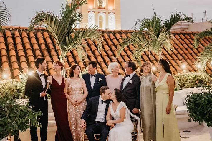 Novios sentados besándose y sus familiares detrás de ellos